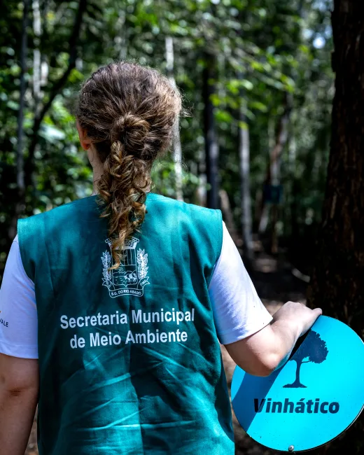 Monitora da Secretaria de Meio Ambiente na trilha, ao lado da placa da árvore vinhático